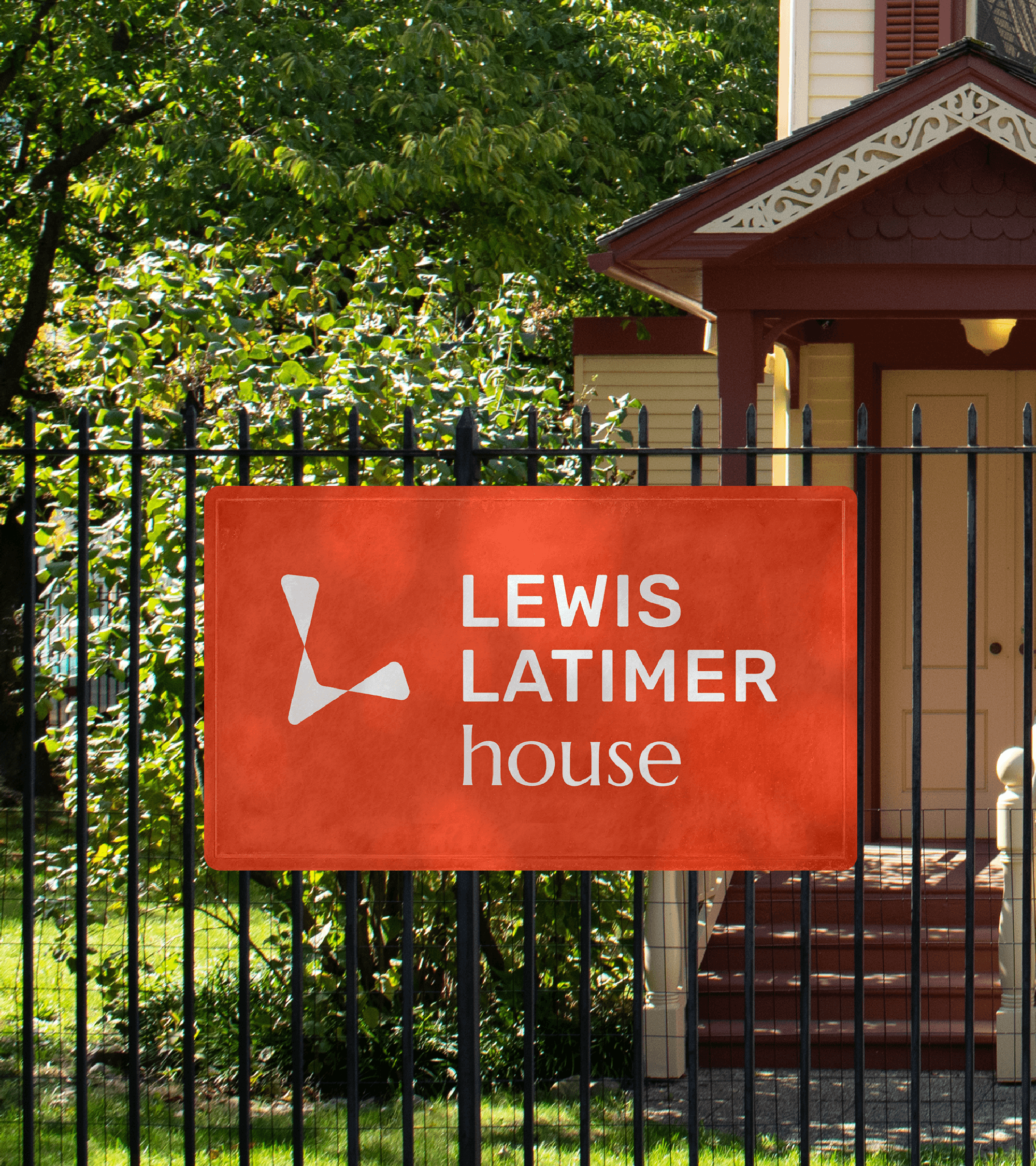 House with greenery behind iron fence. Orange banner with Lewis Latimer house logo displaying on fence.