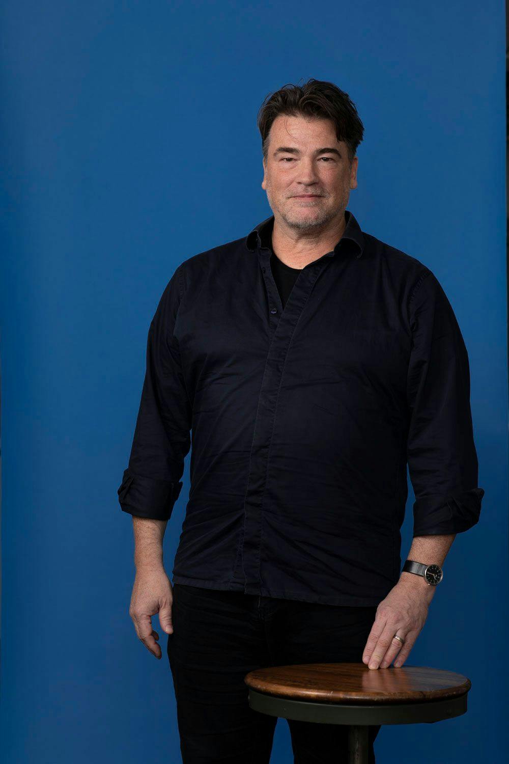 Mark Kingsley standing behind a stool against a blue backdrop.