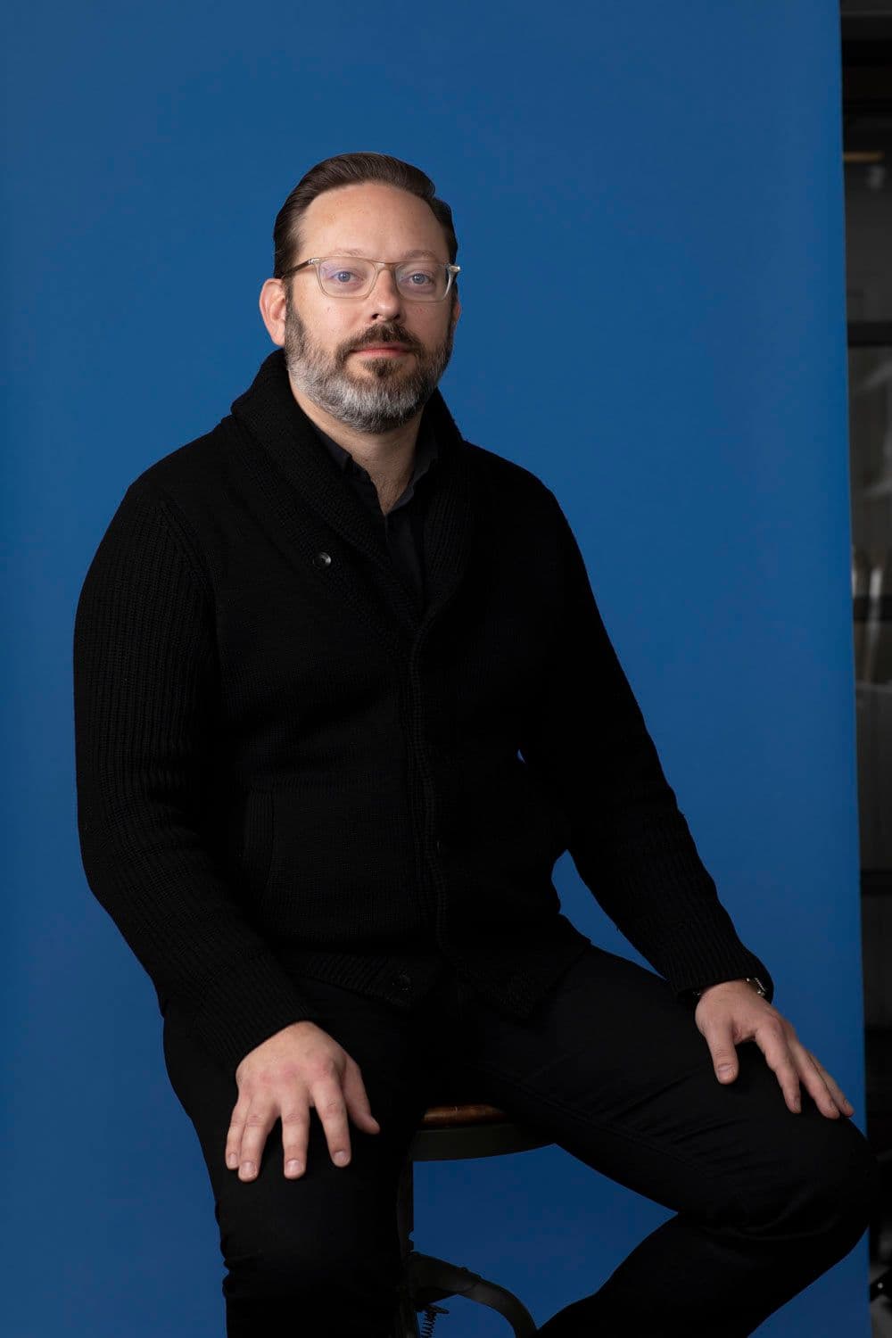 Tosh Hall sitting on a stool against a blue backdrop.